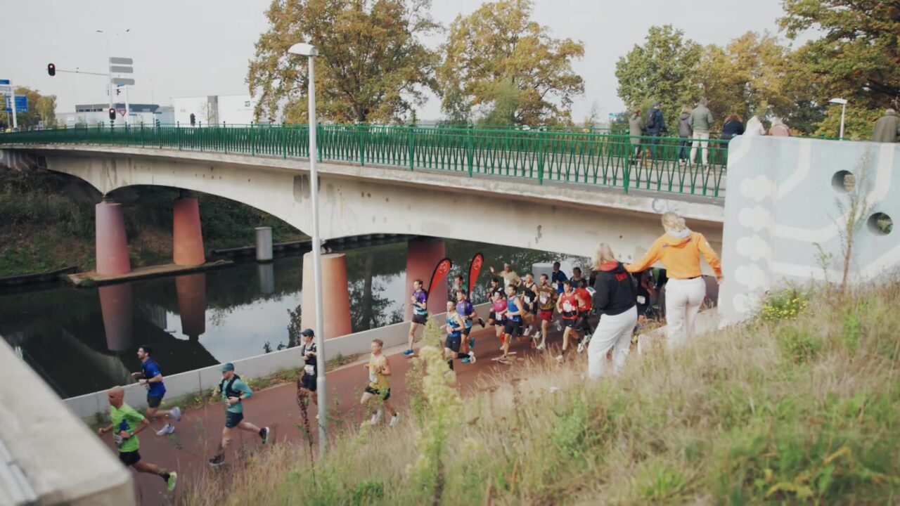 Lopers van de marathon van Eindhoven langs het water onder een brug, met publiek op de oever