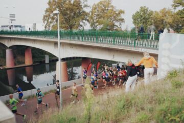 Lopers van de marathon van Eindhoven langs het water onder een brug, met publiek op de oever